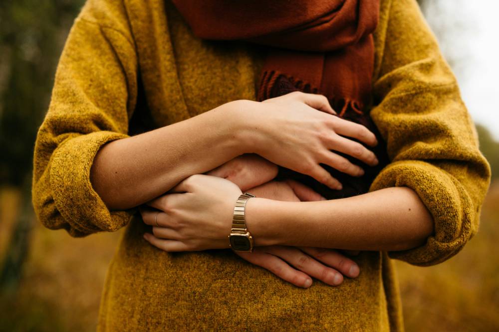 Person in mustard colored sweater and rust colored scarf holding a person's arms and hands